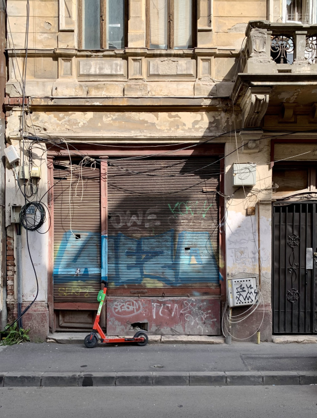 Ein roter Roller, der auf einer Straße neben einem Gebäude mit Fenstern, Rollläden und einem Tor geparkt ist. Sichtbare Drähte und Gegenstände entlang der Straße und Text am unteren Rand.