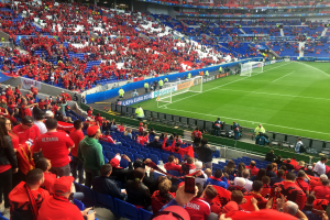 Ein volles Stadion mit Zuschauern, einige sitzen und einige stehen, schauen ein Fußballspiel. Banner mit Text sind sichtbar, zusammen mit einem Torpfosten und einem Dach mit Deckenbeleuchtung.