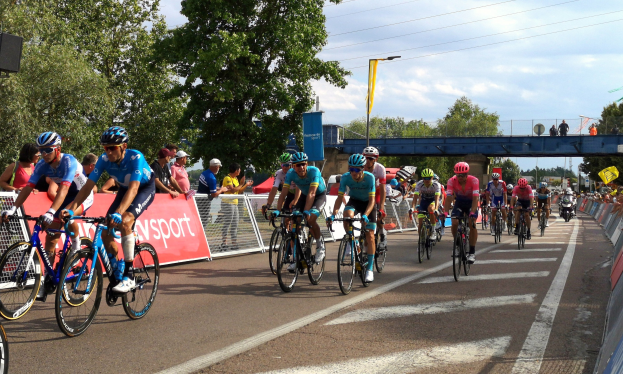 Gruppe von Radfahrern mit Helmen, Brillen und Handschuhen auf einer Straße mit Geländern, Bannern, Bäumen, einer Brücke, Laternen, Drähten und einem bewölkten Himmel im Hintergrund.