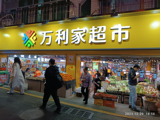 Eine Gruppe von Menschen geht an einem chinesischen Supermarkt in Hong Kong vorbei, mit sichtbaren Gemüse, Früchten und Schildern im Inneren des Ladens.