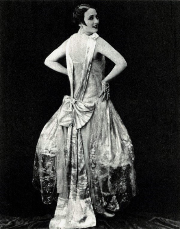 Eine Frau in einem Kleid im 1920er-Stil mit hohem Kragen, langen Ärmeln und einem weiten Rock steht selbstbewusst in einem Schwarz-Weiß-Foto, ihr Haar ist elegant frisiert und das Make-up dezent natürlich.