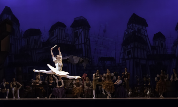Eine Ballerina in einem weißen Tutu und Spitzenschuhen tanzt auf der Bühne, während das Publikum zusieht, mit einem Burgenbild im Hintergrund.