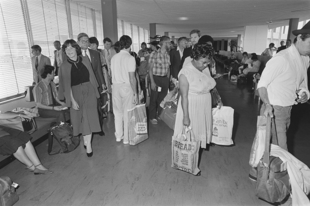 Menschen gehen durch ein schwarz-weißes Flughafen-Terminal mit Gepäck, einige sitzen auf Bänken und andere stehen, Fenster mit Jalousien links, ein Mann und eine Frau stehen nebeneinander und halten Taschen.