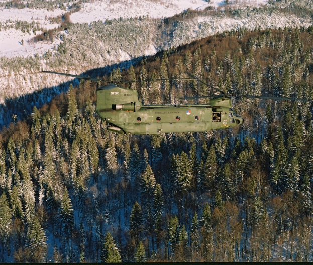 Militärhubschrauber fliegt über einen schneebedeckten Wald mit Bergen im Hintergrund, Text unten sichtbar.