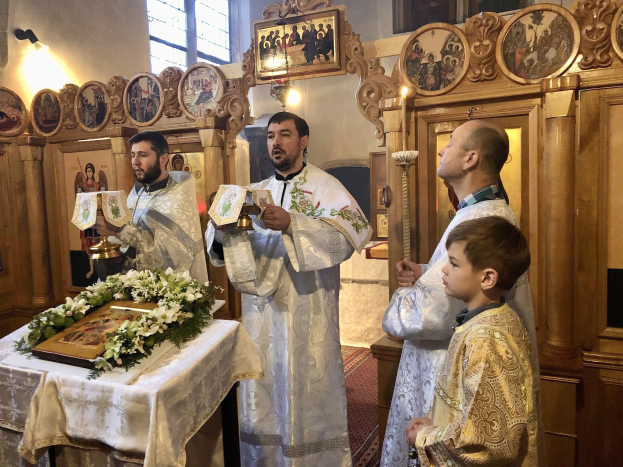 Eine Gruppe von Menschen steht um einen Tisch in einer Kirche, hält Bücher in den Händen, mit einem Tuch und einem Blumenstrauß auf dem Tisch und Foto-Rahmen, Lichtern, einem Fenster und einem hölzernen Gegenstand im Hintergrund.