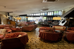 Ein geräumiger Eingangsbereich mit zahlreichen Stühlen, Tischen, Sofas mit Kissen, Wandlampen, einer Deckenklimaanlage und großen Glasfenstern an der Rückseite.