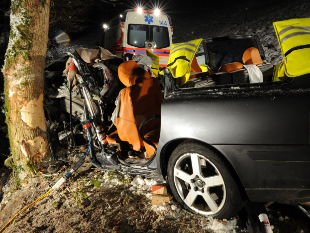 Ein beschädigtwagen am Rande einer verschneiten Straße mit einem Krankenwagen im Hintergrund, umgeben von Bäumen.