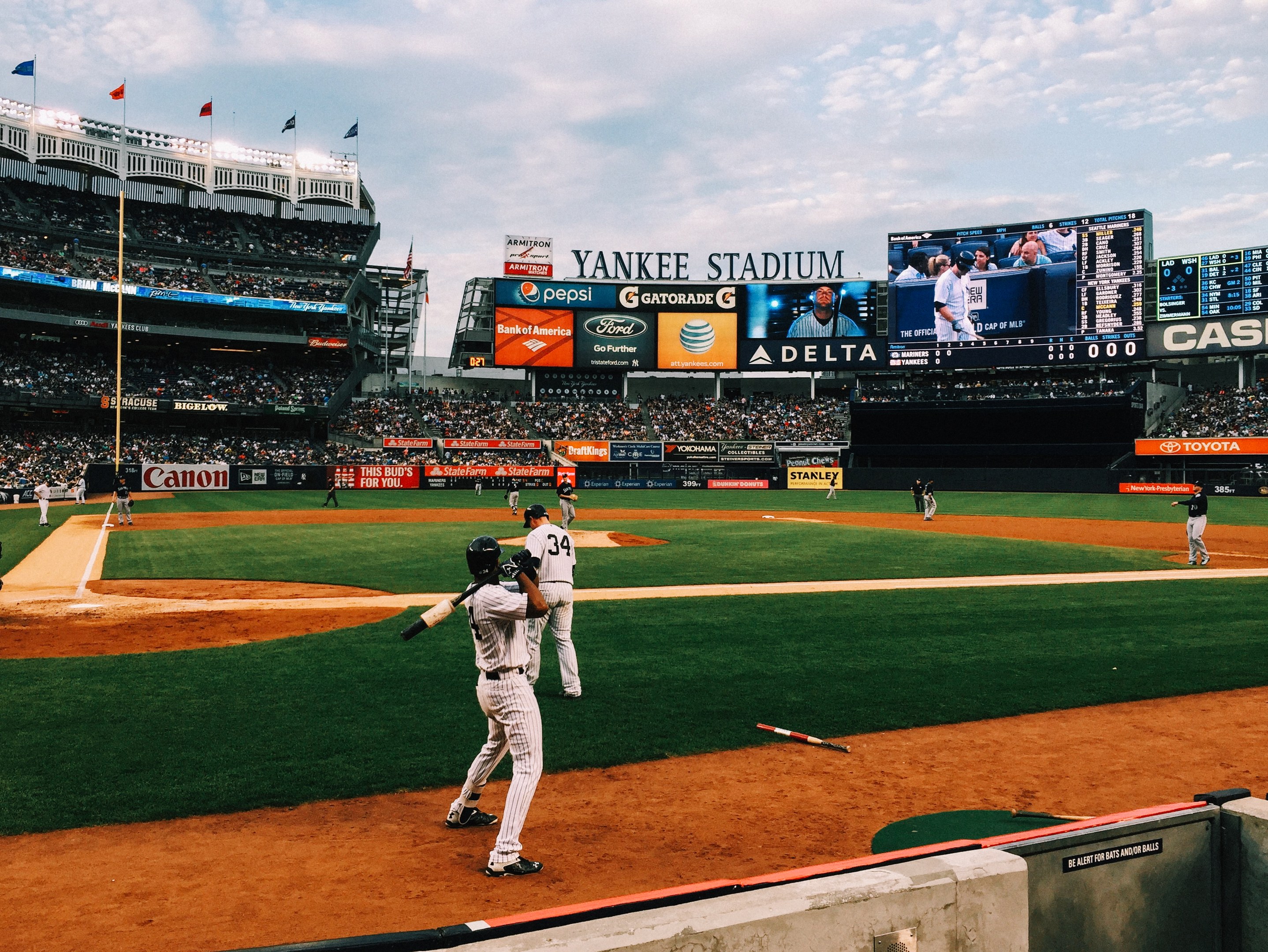 Ein Baseball-Spiel im Yankee Stadium, mit Spielern und Zuschauern, umgeben von Stadion-Features wie Zäunen, Fahnen, Anzeigetafeln, einem Display und Deckenlampen unter einem bewölktem Himmel.
