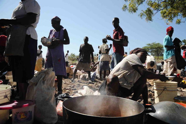 Eine Gruppe von Menschen, die in l├Ąndlicher Umgebung Essen zubereiten, mit Eimern und Abdeckungen auf dem Boden und B├Ąumen im Hintergrund.