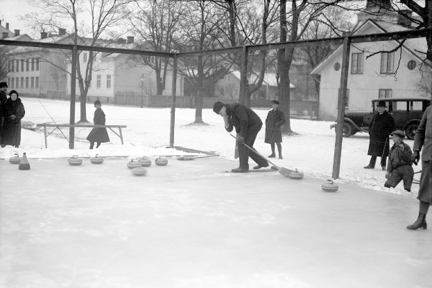Schwarzes Bild einer Gruppe von Menschen, die Curling auf einem Eisplatz spielen, der von einem Zaun umgeben ist, mit einer Bank, Bäumen, Gebäuden und einem geparkten Fahrzeug im Hintergrund.