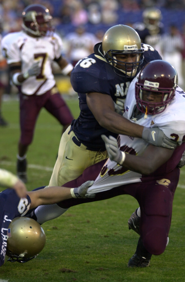 Zwei Fußballspieler in Helmen und Uniformen ringen auf dem Feld, während andere zusehen.