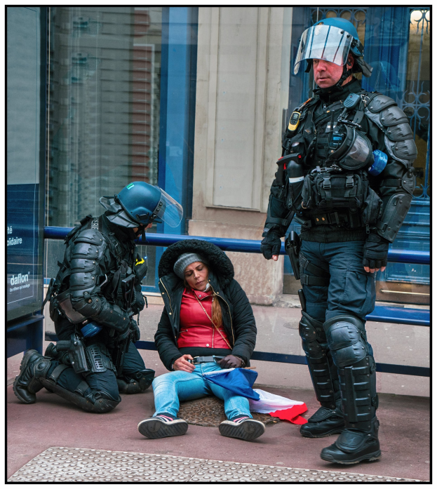 Eine Frau sitzt auf dem Boden neben zwei uniformierten Polizeibeamten, mit einem Gebäude mit Glastüren und einem Schild im Hintergrund.