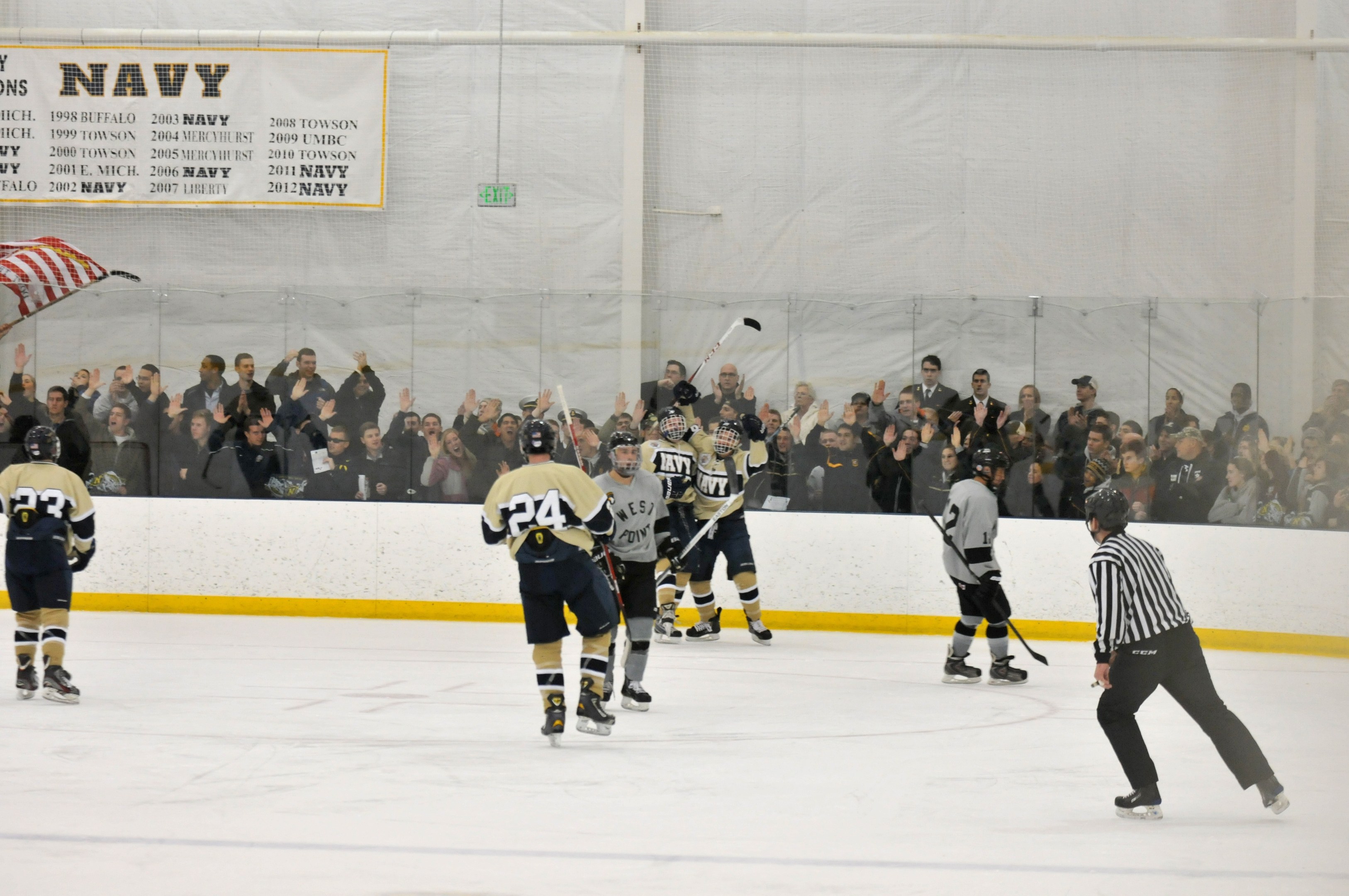 Gruppe von Menschen, die Eis-hockey auf einem Eisstadion spielen, mit Zuschauern auf den Tribünen, die Helme tragen und Hockey-Schläger halten, mit einer Wand im Hintergrund und einer Fahne links.