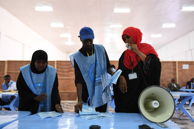 Eine Gruppe von Menschen versammelt sich um einen Tisch mit einem Megafon, Papieren und anderen Gegenständen an einem Ort, der wie eine Wahllokalität aussieht, während andere im Hintergrund sitzen und stehen.