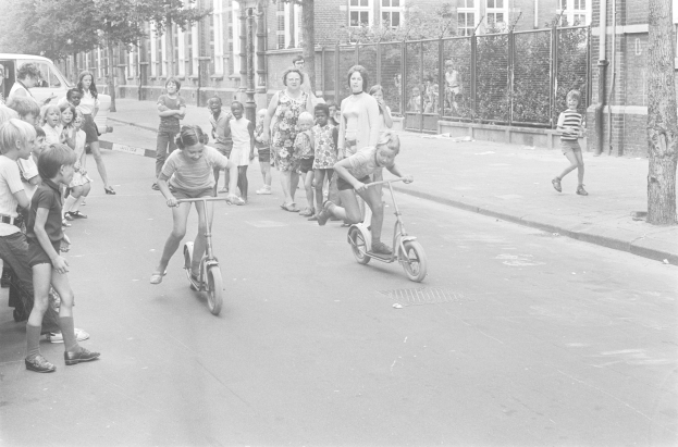 Schwarz-weißes Bild von Kindern, die auf Roller die Straße hinunterfahren, mit einem geparkten Auto auf der linken Seite, Gebäuden, Bäumen, Polen und einem Zaun im Hintergrund.