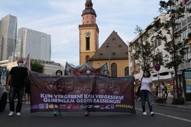 Eine Gruppe von Menschen mit Masken, die eine Straße entlanggehen und ein Banner halten, mit einem geparkten Auto auf der linken Seite, Gebäuden und einem Kirchturm im Hintergrund unter einem klaren blauen Himmel.