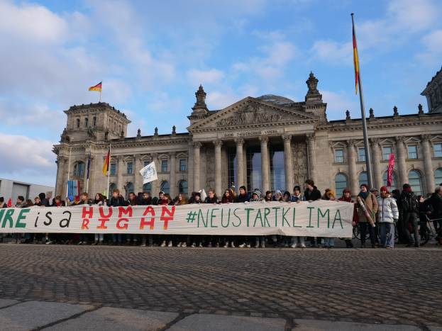 Gruppe von Menschen vor dem Reichstaggebäude in Berlin, Deutschland, mit einem Banner mit der Aufschrift "Wir sind ein Menschenrecht", das architektonische Details des Gebäudes und Flaggen im Hintergrund zeigt.
