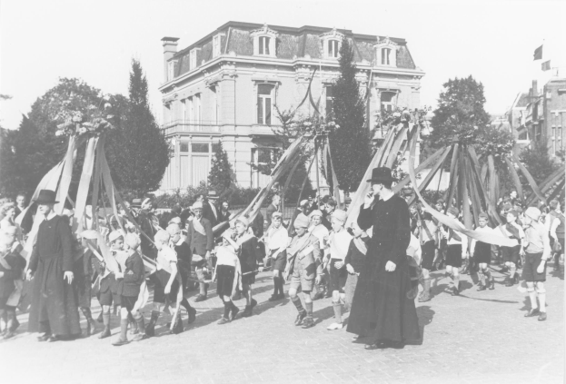 Schwarz-weißes Bild einer Gruppe von Menschen, die eine Straße entlanggehen, einige halten Fahnen, mit Hüten, Bäumen, Gebäuden und einem sichtbaren Himmel im Hintergrund, das einen Maibaum als Zeichen für ein Maifest zeigt.