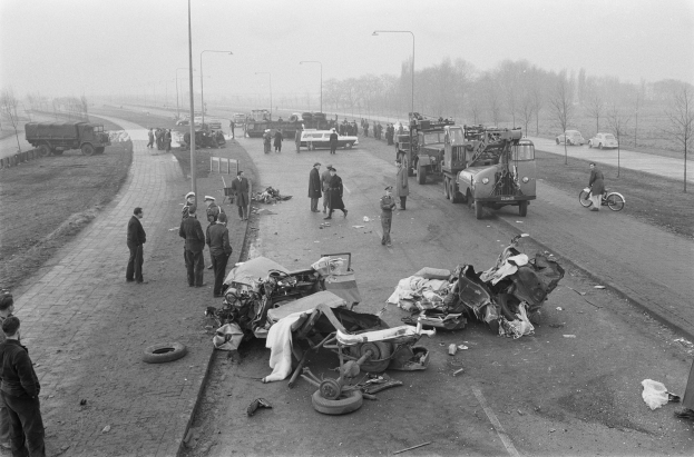 Schwarz-weiß-Szene eines Autounfalls am Straßenrand mit mehreren Fahrzeugen, einer Gruppe von Menschen in der Nähe und Laternen, Bäumen und Himmel im Hintergrund.