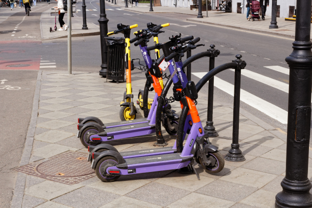 Eine Reihe von Elektro-Scootern, die auf der Straße geparkt sind, mit Menschen, die auf dem Gehweg gehen und Fahrräder fahren, einem Müllcontainer im Vordergrund und Gebäuden, Bäumen und Pflanzen im Hintergrund.