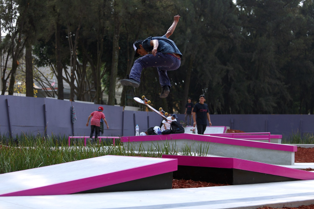 Ein Skateboarder zeigt eine Luftfigur auf einer Rampe, während eine Gruppe von Menschen von unten zuschaut, einige halten Skateboards, mit verstreuten Taschen, Flaschen und anderen Gegenständen in der Nähe.