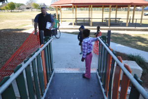 Eine Gruppe von Menschen auf einem Spielplatz, mit einem Mann, der ein Buch hält und ein Kind im Rollstuhl, umgeben von einem Zaun, Gras, Pflanzen, einem Weg, Bänken, einem Schuppen, einem Auto, Pfählen, Bäumen, Häusern und einem bewölkten Himmel.