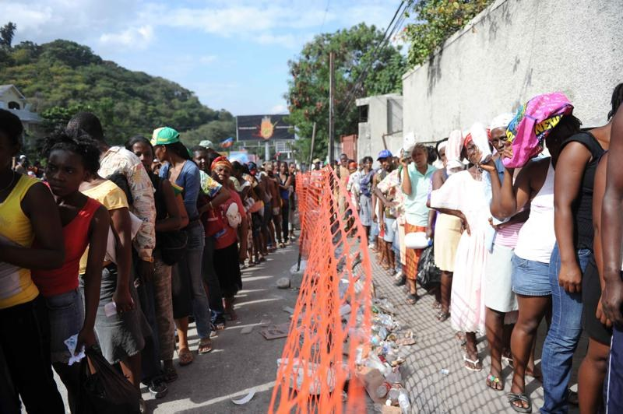 Lange Schlange von Menschen, die vor einem Zaun auf ihre Abstimmung warten, mit einer Wand, Bäumen, Gebäuden, Strommasten und einer Werbetafel im Hintergrund.