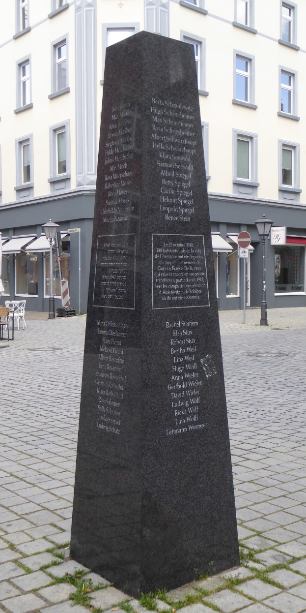 Ein Denkmal mit Inschrift steht auf einem Stadtplatz umgeben von Gebäuden, Straßenmöblierung und Menschen unter einem sichtbaren Himmel.