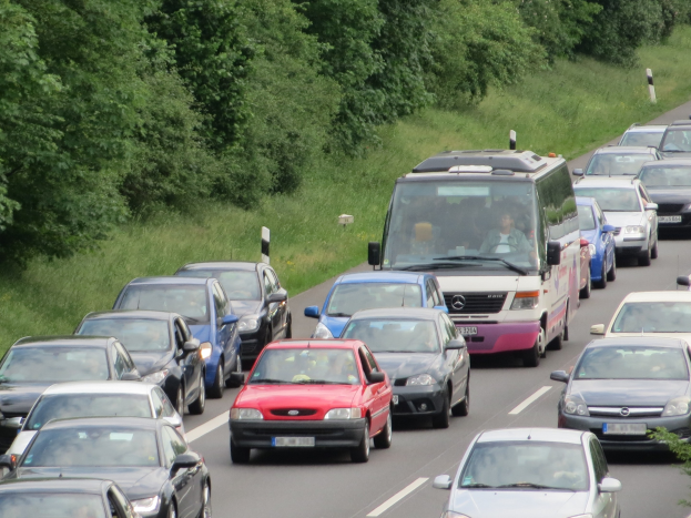 Verkehrsstau auf einer Autobahn mit zahlreichen Autos und einem Lieferwagen, mit Menschen in den Fahrzeugen und Bäumen und Gras im Hintergrund.