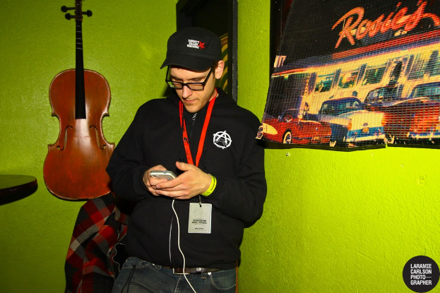 Eine Person mit einer Mütze und einem Ausweis hält ein Mobiltelefon in der Hand und schaut auf dessen Bildschirm, mit einer Gitarre links daneben und einem Plakat rechts.