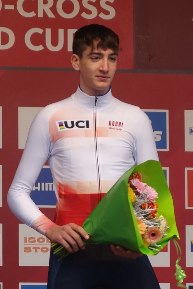 Ein Mann steht auf einem Podium und hält einen Blumenstrauß, feiernd mit einem selbstbewussten und stolzen Ausdruck.