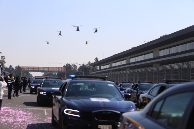 Eine Gruppe von Polizeifahrzeugen vor einem Gebäude geparkt, mit Menschen auf der linken Seite der Straße; im Hintergrund sind Bäume, Pfosten, eine Brücke und ein klarer blauer Himmel zu sehen, mit vier Hubschraubern, die oben im Bild fliegen.