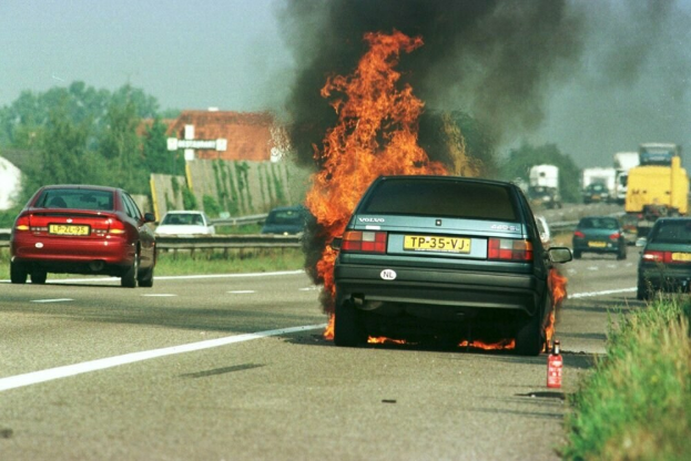 Ein Auto, das in Flammen steht, auf der Seite der Straße, umgeben von anderen Fahrzeugen, mit Bäumen, Gebäuden und einem klaren blauen Himmel im Hintergrund, Gras auf der rechten Seite und ein Feuerlöscher in der Nähe.