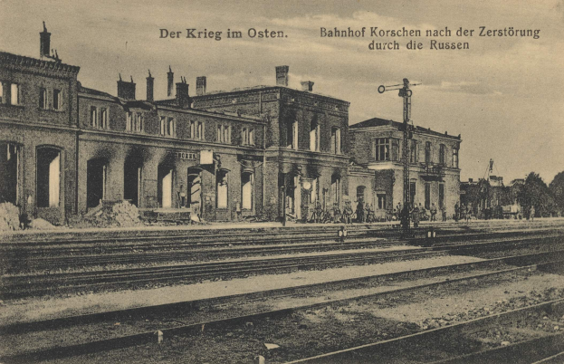 Schwarzes und weißes Foto des Bahnhofs Korschen in Ostern, Deutschland, das Gleise, Bahnhofsgebäude, Menschen, Bäume und einen Himmel mit dem Text "Bahnhof Korschen in Ostern, Deutschland" oben und unten zeigt.