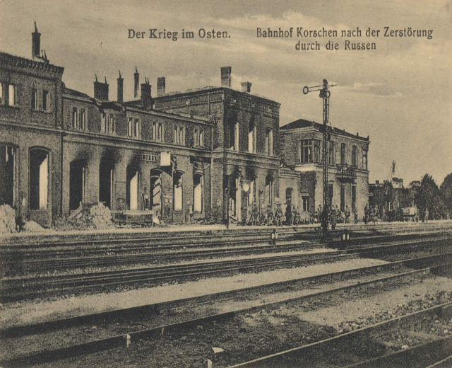 Schwarzes und weißes Foto des Bahnhofs Korschen in Ostern, Deutschland, das Gleise, Bahnhofsgebäude, Menschen, Bäume und einen Himmel mit dem Text "Bahnhof Korschen in Ostern, Deutschland" oben und unten zeigt.