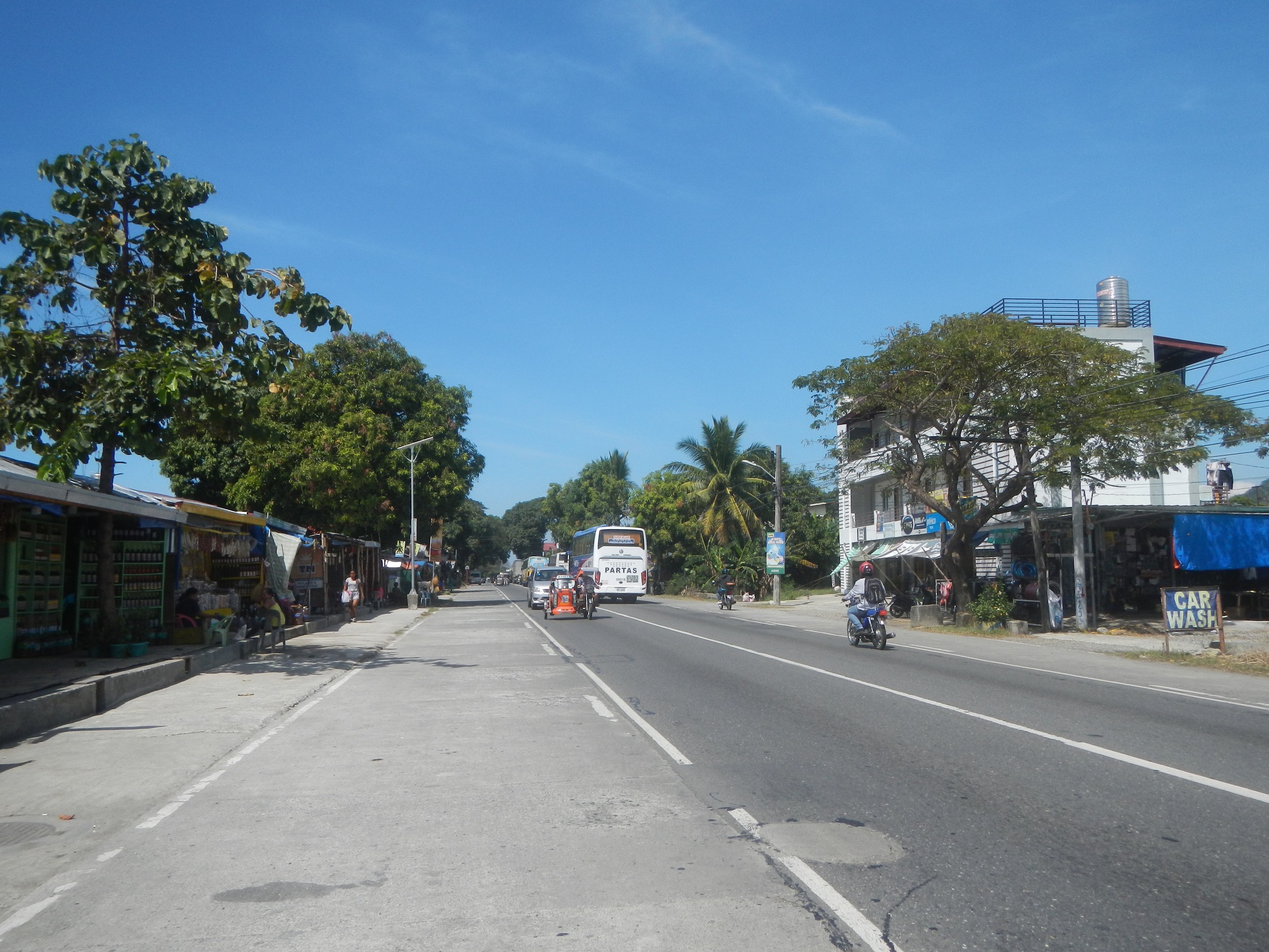 Stadtstraße mit fahrenden Fahrzeugen, Fußgängern auf dem Gehweg, Geschäftsfronten links, Bäumen, Gebäuden, Strommasten mit Drähten und einem bewölkten Himmel.