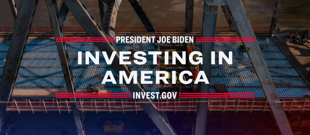 Präsident Joe Biden in der Nähe einer Brücke mit dem Text "In Amerika investieren" im Vordergrund, umgeben von verstreuten Gegenständen.