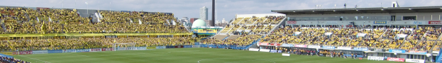 Ein großes Stadion mit Zuschauern bei einem Fußballspiel auf saftigem Grün, umgeben von Werbetafeln, mit Gebäuden und Pfosten im Hintergrund unter einem klaren blauen Himmel.