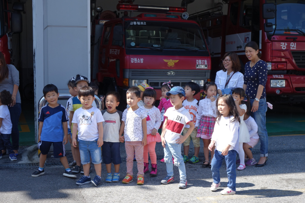 Eine Gruppe von Kindern vor einem Feuerwehrauto auf einer Feuerwache, einige tragen Mützen, mit weiteren Feuerwehrautos im Hintergrund.