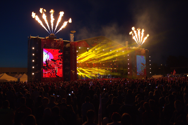 Eine große Menschenmenge auf einem Musikfestival schaut auf einer hell beleuchteten Bühne performende Künstler, mit Zelten links und Feuerwerk am dunklen Himmel dahinter.