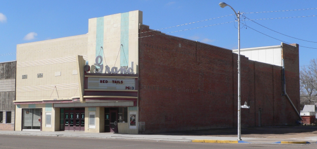 Ein altes Kino an einer Straßenecke, umgeben von Gebäuden, Straßenlaternen, Strommasten, Stromkabeln, Fahrzeugen, Bäumen und einem bewölkten Himmel.