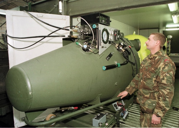 Ein Mann in militärischer Uniform steht neben einer großen grünen Bombe in einem unordentlichen industriellen Setting mit Maschinen, Drähten und einer beleuchteten Hintergrundwand, mit Text unten.