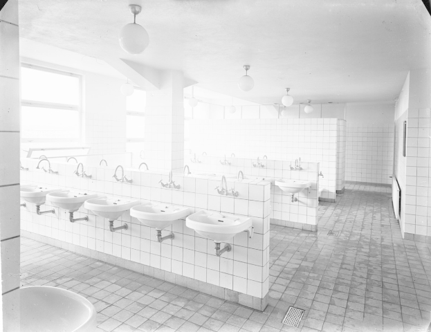 Schwarzes und weißes Foto einer öffentlichen Toilette mit Waschbecken, Hähnen, Wänden, Lampen und Fenstern.