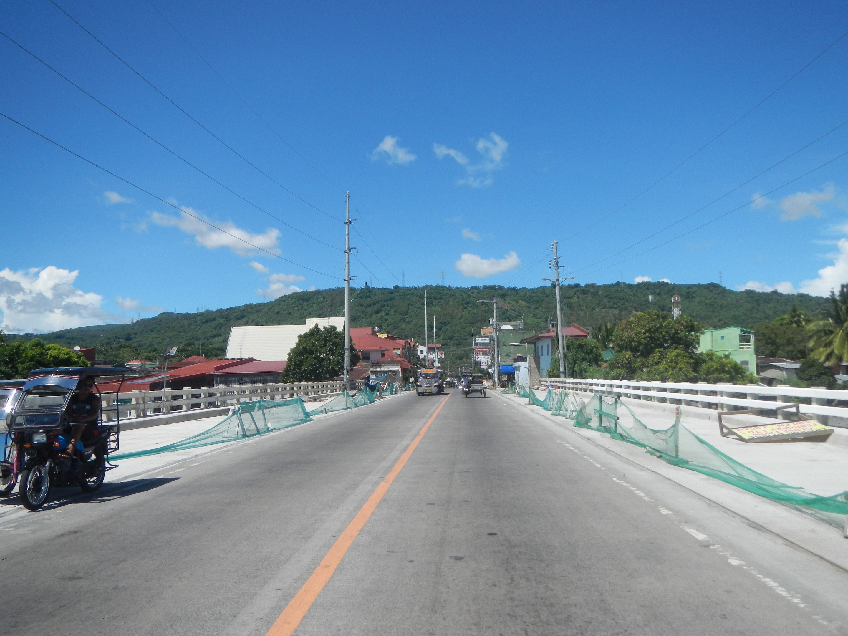 Autobahn mit fahrenden Fahrzeugen, ein geparktes Motorrad, Geländer, Strommasten mit Drähten, Bäume, Gebäude, Hügel und einen bewölkten Himmel im Hintergrund.