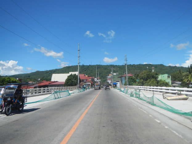 Autobahn mit fahrenden Fahrzeugen, ein geparktes Motorrad, Geländer, Strommasten mit Drähten, Bäume, Gebäude, Hügel und einen bewölkten Himmel im Hintergrund.