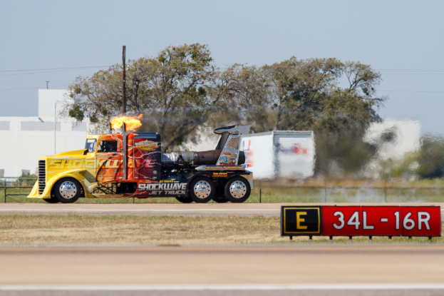 Ein Lkw mit Flammen fährt eine Straße mit Gras, einem Zaun, Bäumen, Polen, Drähten, einem Gebäude und einem Himmel im Hintergrund entlang.