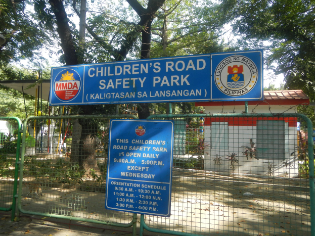 Metallzaun um einen Kinderverkehrssicherheitspark mit zwei blauen Informationsschildern, einem kleinen Überdachten Gebäude, Pflanzen, Bäumen und einem bewölktem Himmel.