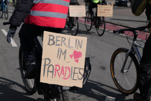 Eine Gruppe von Menschen, die auf Fahrrädern eine Straße entlangfahren, mit einem "Berlin I'm Paradies"-Schild im Vordergrund und einem unscharfen Auto im Hintergrund.