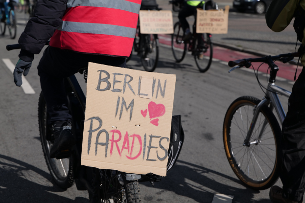 Eine Gruppe von Menschen, die auf Fahrrädern eine Straße entlangfahren, mit einem "Berlin I'm Paradies"-Schild im Vordergrund und einem unscharfen Auto im Hintergrund.