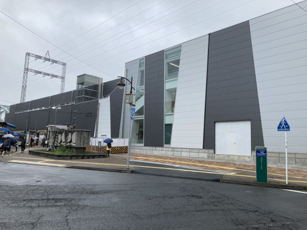 Großes modernes Gebäude mit zahlreichen Fenstern, identifiziert als Tokyo Metropolitan Government Building, umgeben von Straßeninfrastruktur, Fußgängern mit Regenschirmen, Fahrzeugen, Bäumen und einem bewölkten Himmel.